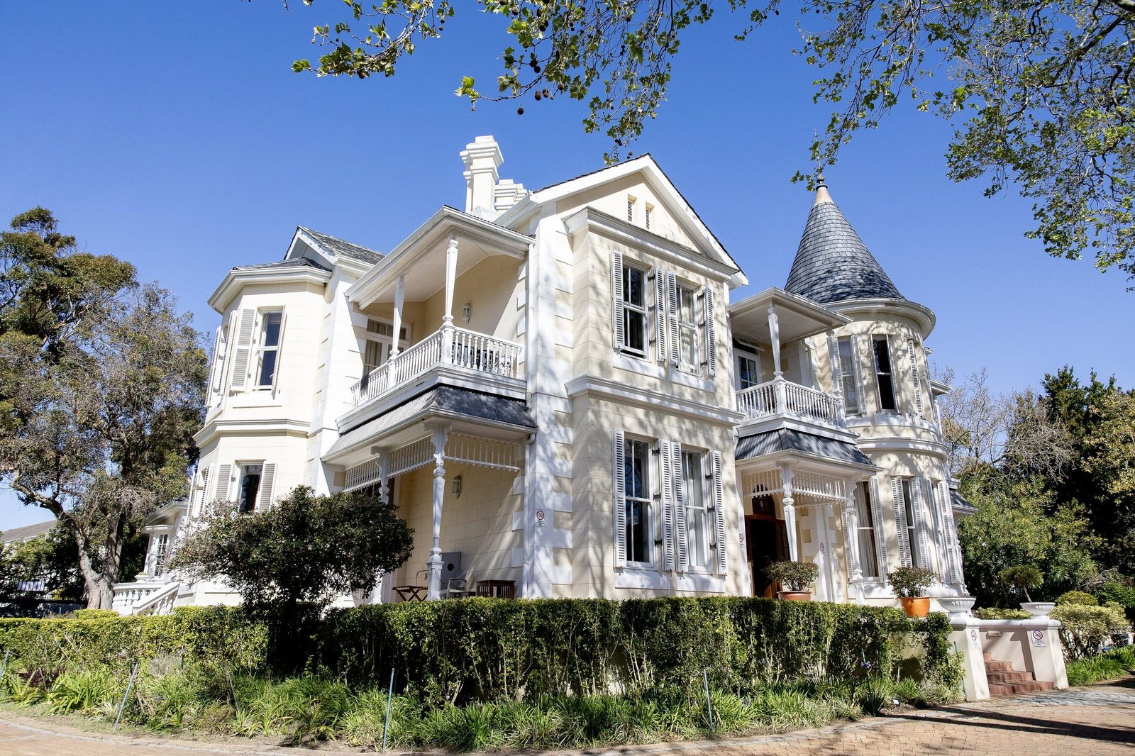 Victoria Haus from Oak Avenue: a white Victorian house with a slate turret, deep verandas and shuttered sash windows, framed by oak branches against a blue sky.