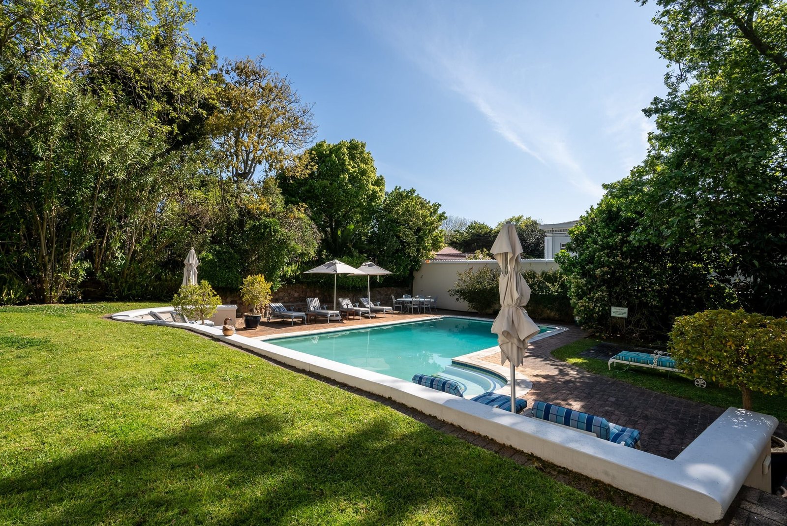 A sunny lawn running down to the pool, with loungers and umbrellas under tall trees.