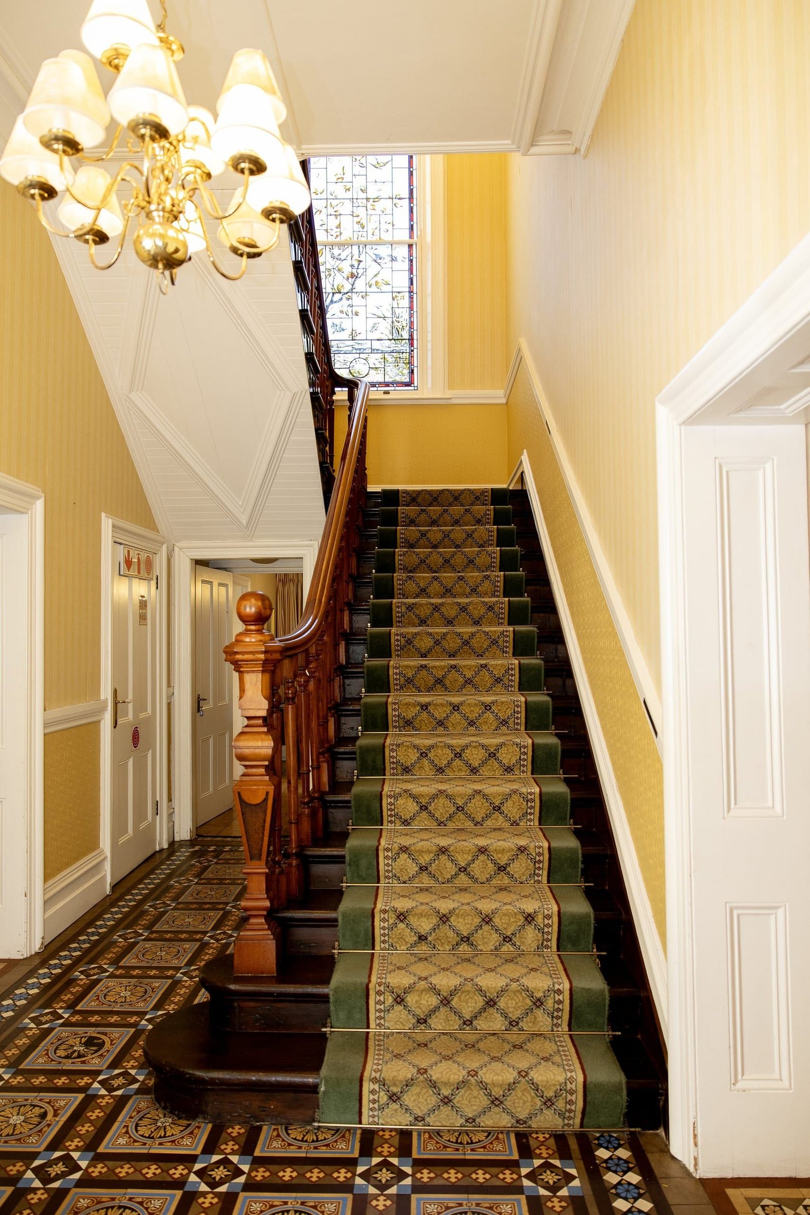 The entrance hall: a brass chandelier, yellow walls, a mahogany banister, a green patterned stair runner and a stained-glass window, over an encaustic tiled floor.