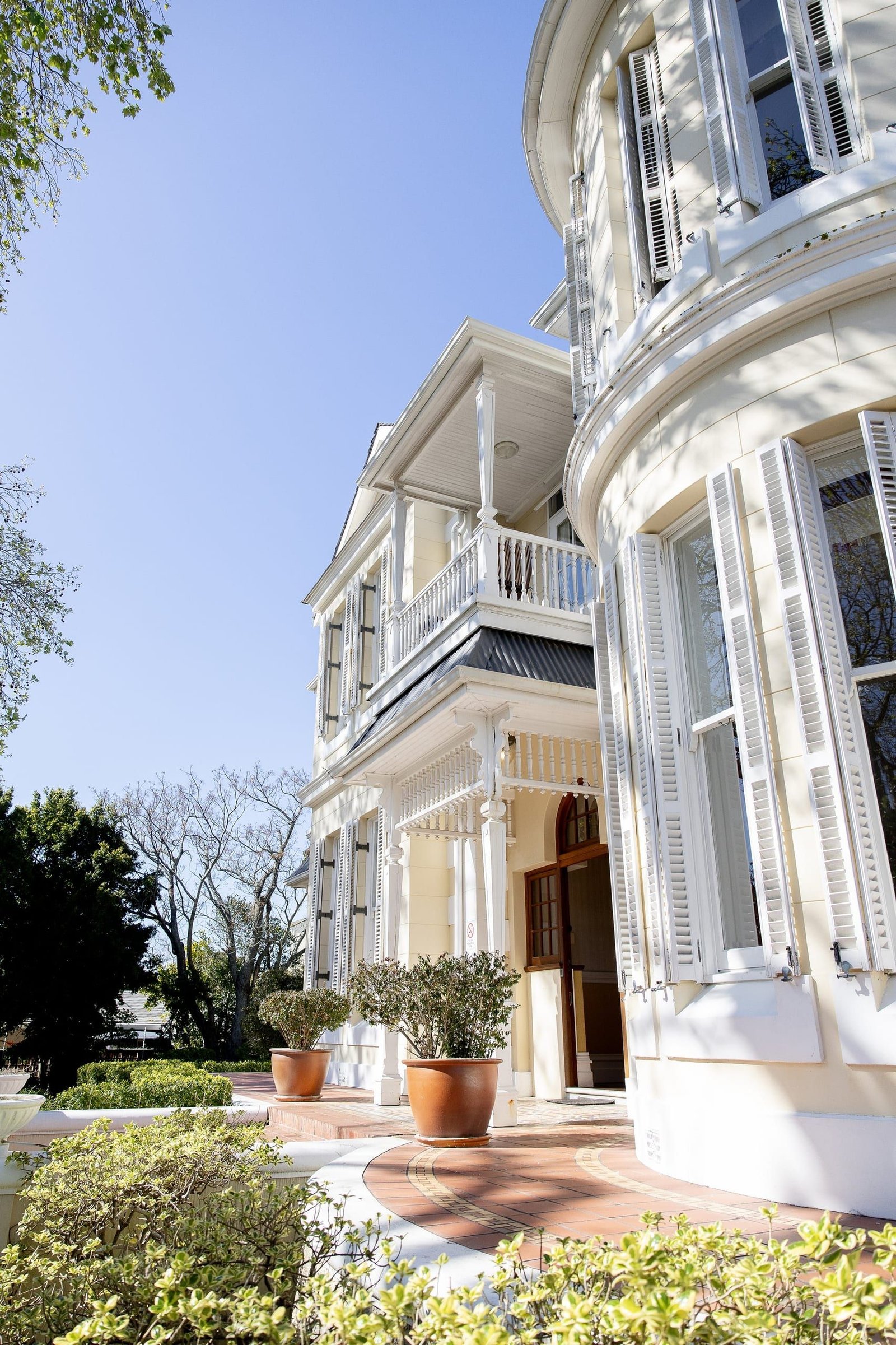 The front entrance beside the curved turret, with white shutters, a veranda above and potted olive trees on a brick path.